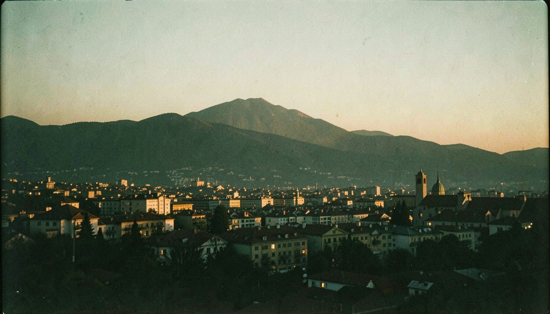 Analogna filmska fotografija panorame grada Tuzle u sumrak s toplim zlatnim svjetlima i siluetom planina u pozadini, miran urbani ugođaj