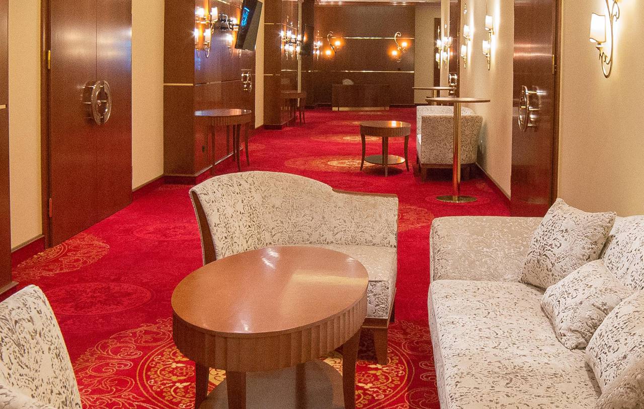 Hotelski hodnik s crvenim tepihom s uzorkom, krem presvučenim klupama, drvenim vratima i zidnim svijećnjacima, topla formalna atmosfera